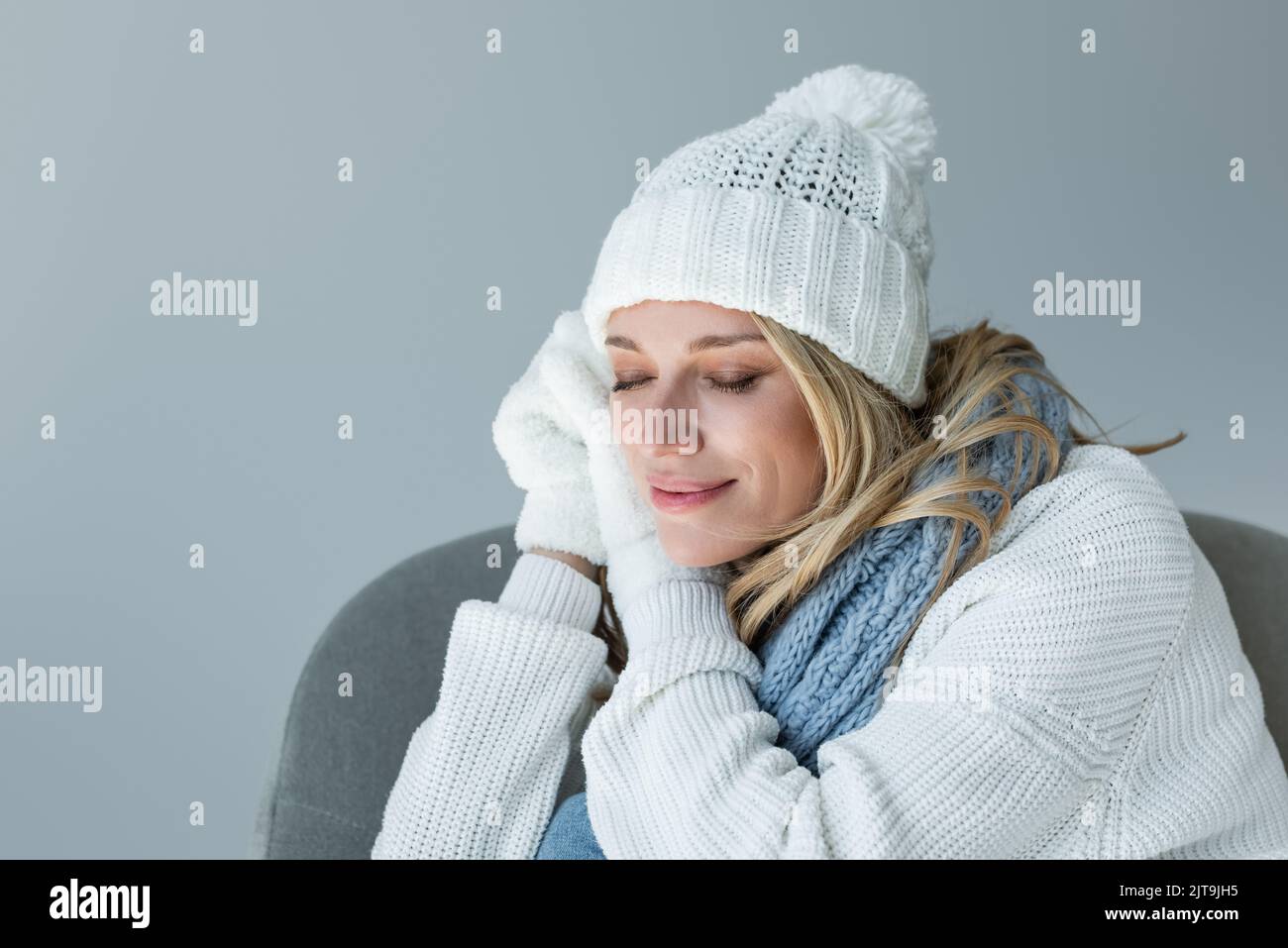 Verschlafte Frau im Winter-Outfit sitzt in einem bequemen Sessel und lächelt isoliert auf grau, Stock Bild Stockfoto
