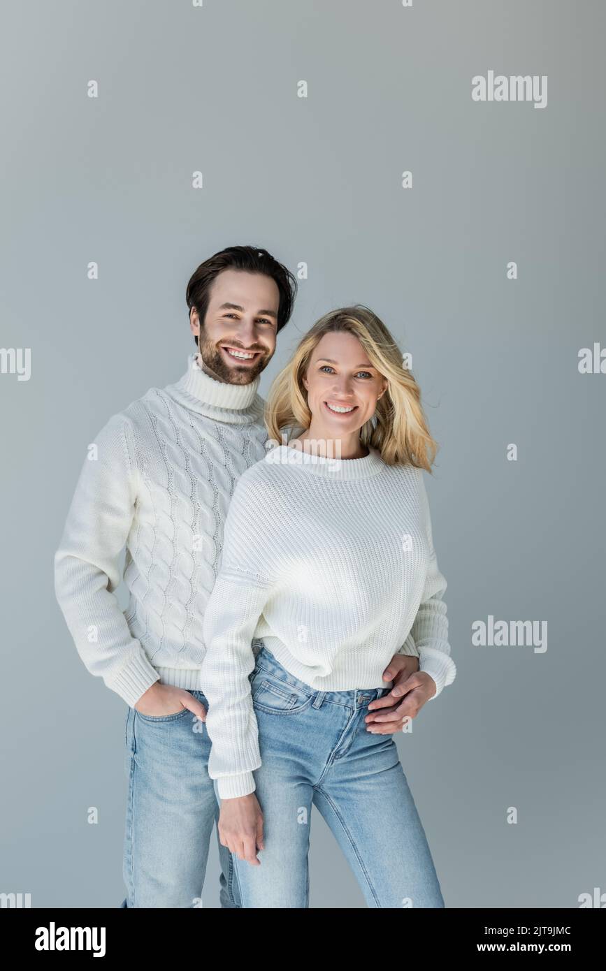 Fröhlicher Mann, der mit der Hand in der Tasche steht und blonde Freundin in einem Pullover umarmt, der auf grauem, Stock-Bild isoliert ist Stockfoto
