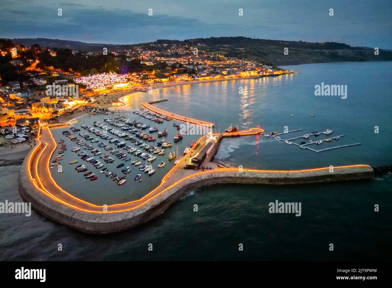 Lyme Regis, Dorset, Großbritannien. 28.. August 2022. Blick aus der Luft, während in der Dämmerung tausende Teelichtkerzen die historischen Cobb-Hafenmauern bei Lyme Regis in Dorset erleuchten. Am Strand befindet sich auch eine riesige beleuchtete Krone zum Gedenken an das Platin-Jubiläum der Königin. Diese spektakuläre Veranstaltung, bei der Menschen eine Kerze in Erinnerung an einen geliebten Menschen sponsern, ist die Geldbeschaffung für Lyme Regis Youth Projects, British Red Cross und Cancer Research UK. Bildnachweis: Graham Hunt/Alamy Live News Stockfoto