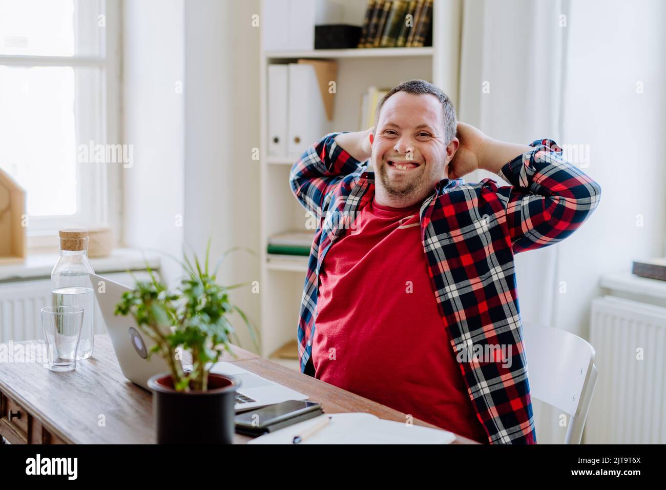 Junger Mann mit Down-Syndrom sitzt am Schreibtisch im Büro und benutzt einen Laptop, schaut auf die Kamera und lächelt. Stockfoto