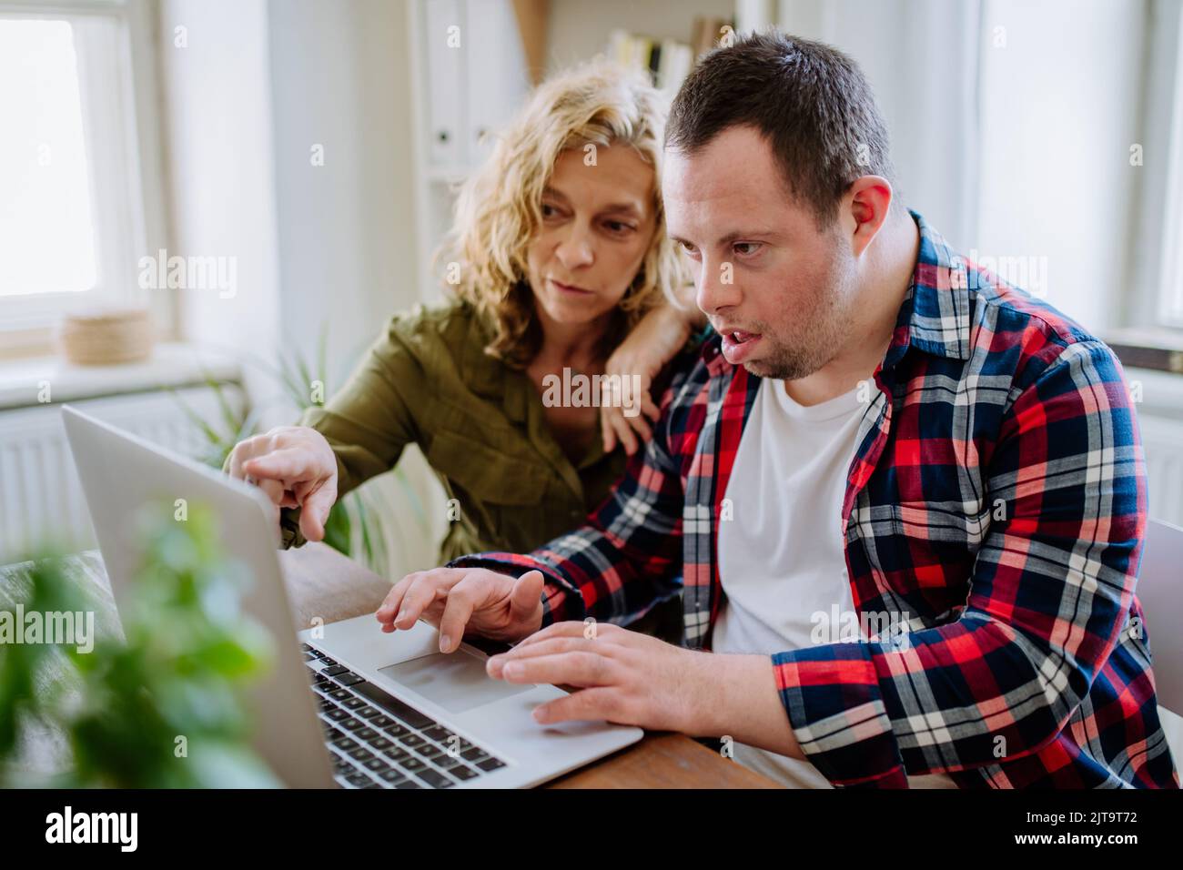 Der junge Mann mit Down-Syndrom sitzt am Schreibtisch im Büro und benutzt einen Laptop, seine Mutter hilft ihm. Stockfoto