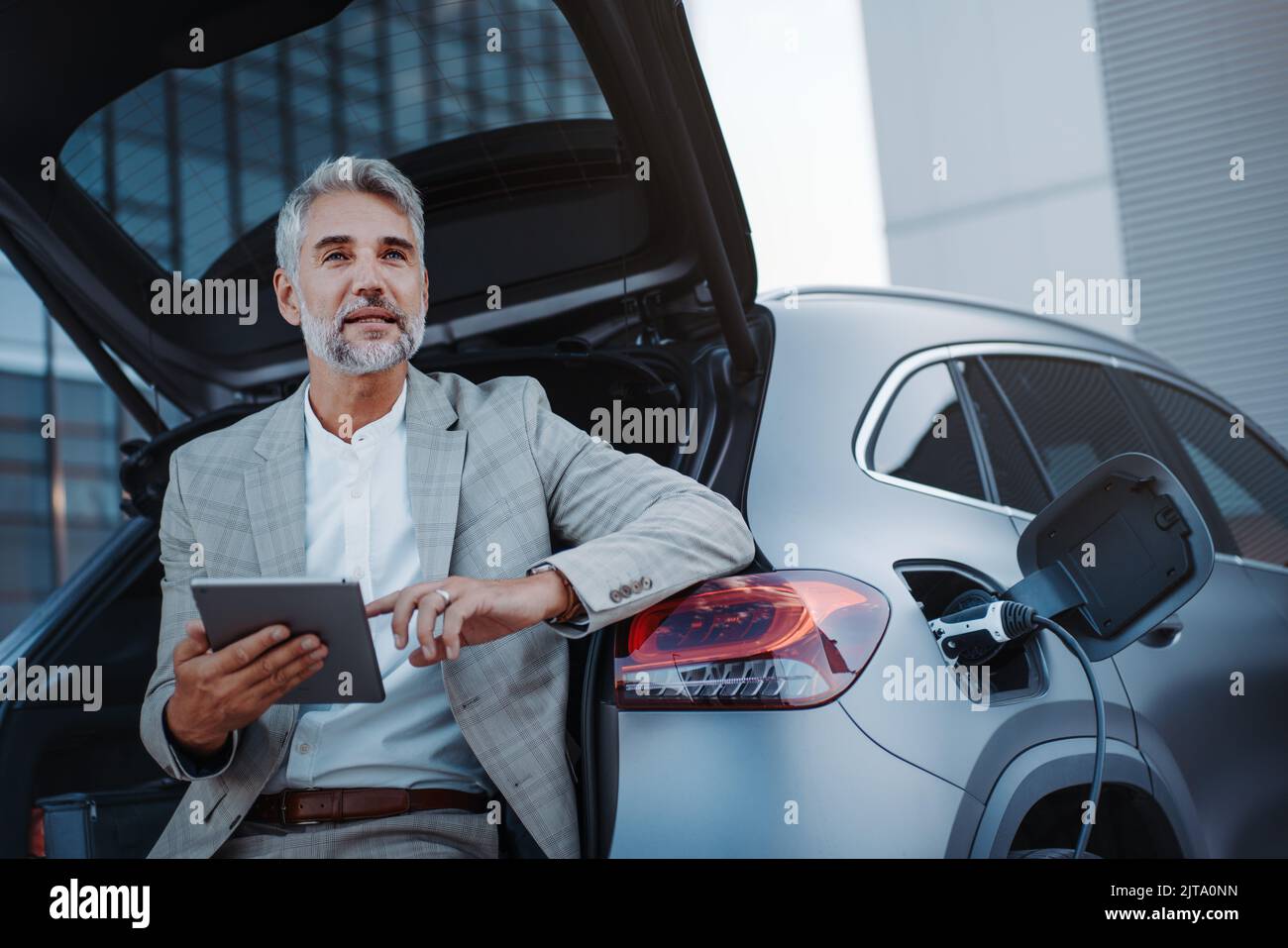 Geschäftsmann mit Tablet, während er das Auto an der Ladestation für Elektrofahrzeuge lädt, Nahaufnahme. Stockfoto