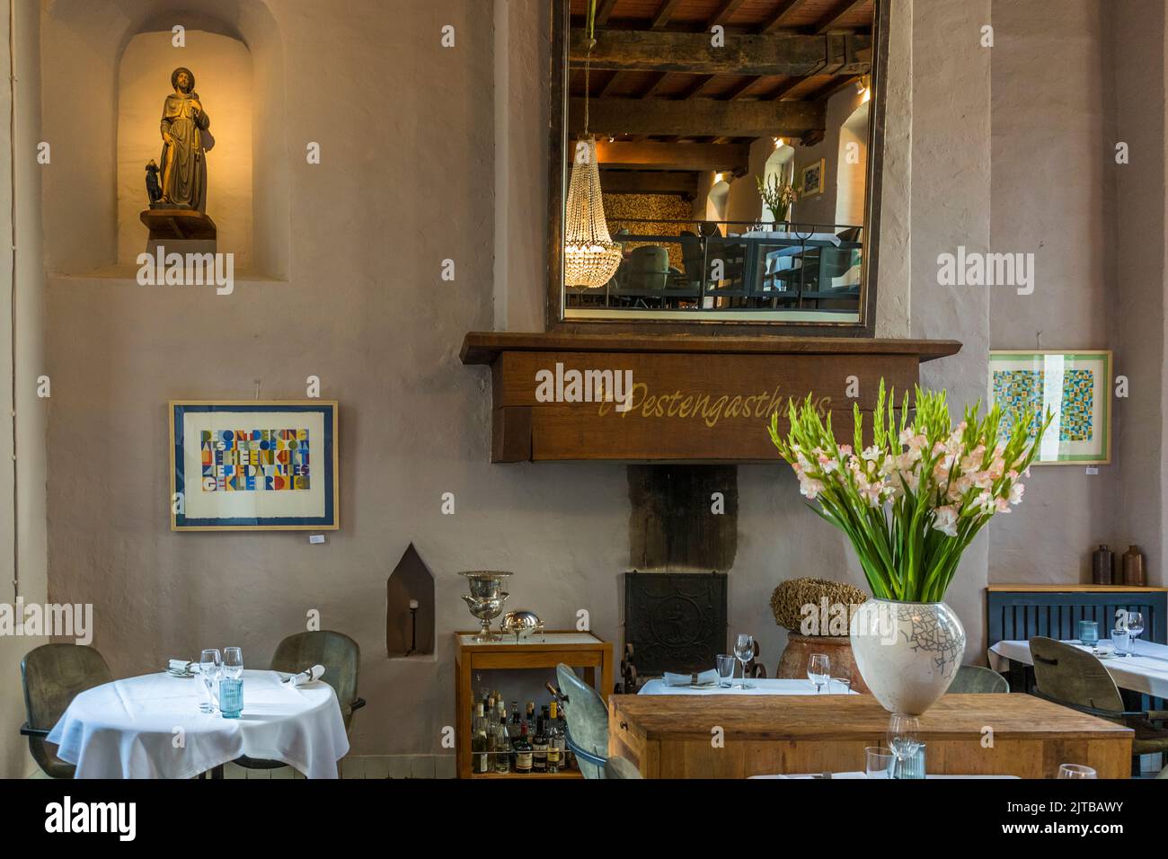 Restaurant 't Pestengasthuys in Zwolle, einer von neun historischen Hansestädten an der IJssel, Provinz Overijssel, Niederlande, begrüßt seine Gäste in den Räumen eines ehemaligen Spitals aus dem 15. Jahrhundert. Ein großer Spiegel hängt über dem Kamin, wobei der Name "'t Pestengasthuys" in den Holzbalken darunter geschnitzt ist, während eine Statue eines Heiligen in einer Wandnische steht. Ein Tisch mit rosa Gladiolen steht im Vordergrund. Stockfoto