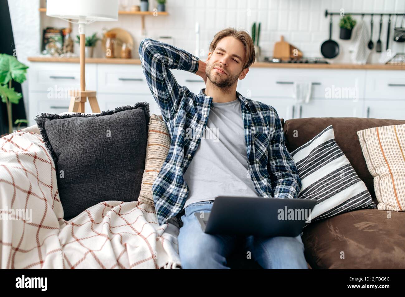 Verärgert kaukasischen stilvollen jungen Mann, sitzt auf dem Sofa im gemütlichen Wohnzimmer, massiert seinen Hals, müde nach einem harten Tag bei der Arbeit und Online-Arbeit, leiden an Gelenkproblemen, eingeklemmt Nerven in den Hals, brauchen Ruhe Stockfoto