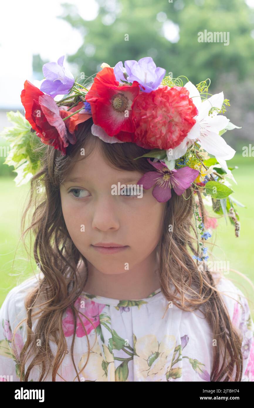Ein kontemplatives Mädchen mit langen Haaren trägt eine Blumenkrone mit roten Mohnblumen, blauer campanula und Blättern. Stockfoto
