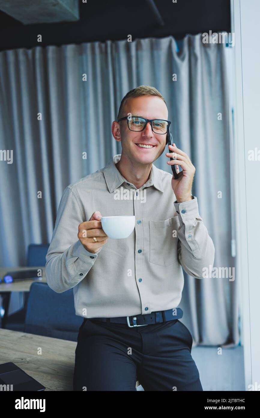 Innenaufnahme des fröhlichen bärtigen männlichen Freiberuflers Arbeit in fernen, verwaltet Finanzen über Smartphone, sitzt am Holztisch, arbeitet mit Dokumentation, hat Stockfoto