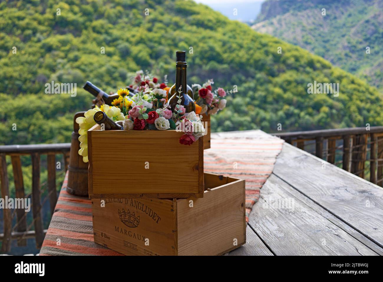 Holzkiste mit leeren Weinflaschen, Früchten und Blumen, Nahaufnahme, flache Tiefe, mit Wald und Bergen im Hintergrund Stockfoto