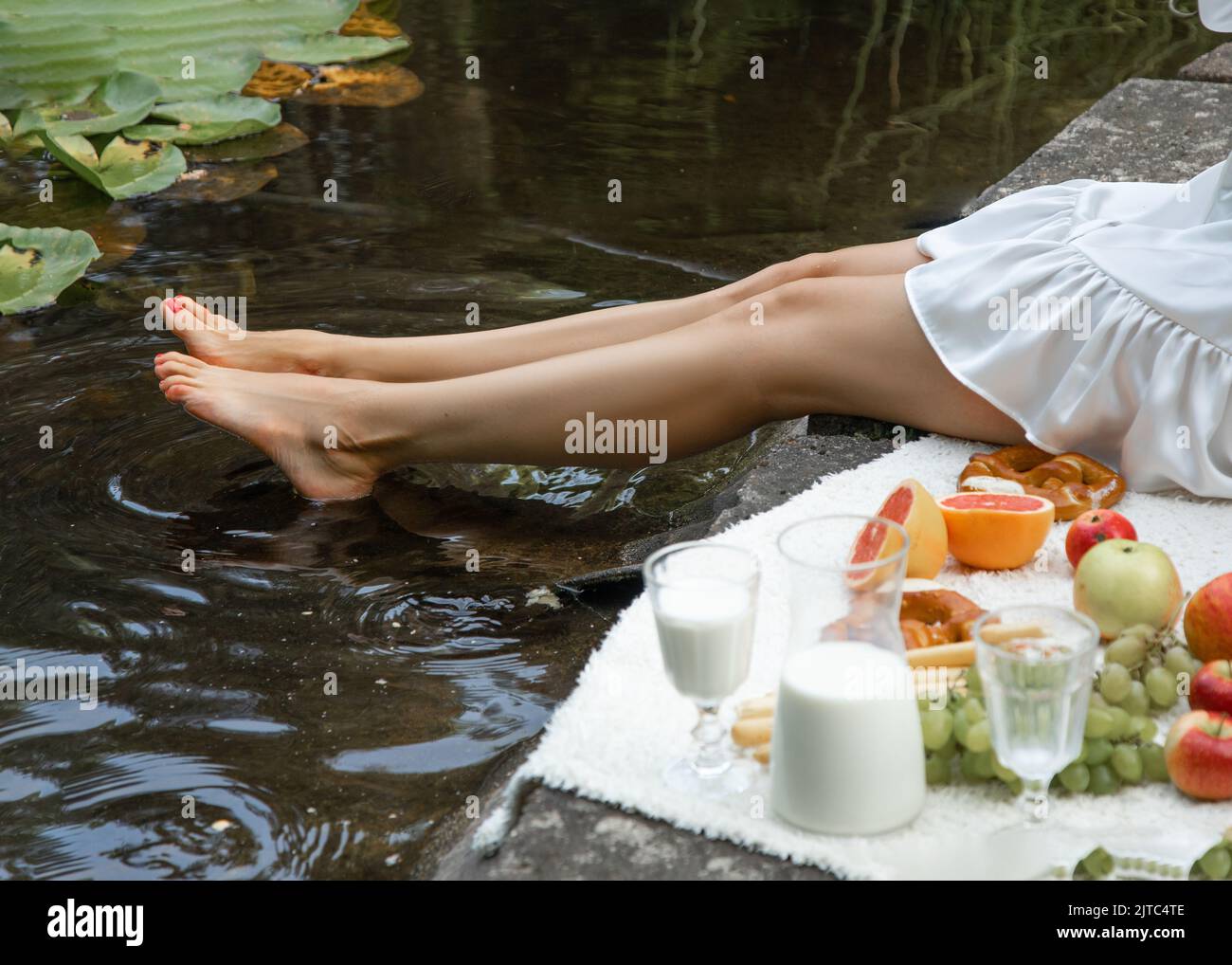 Zugeschnittenes Foto einer jungen Frau, die auf einem karierten mit Früchten, einem Milchkännchen am Rand des Teiches, die Beine über dem Wasser haltend, sitzt. Picknick. Stockfoto