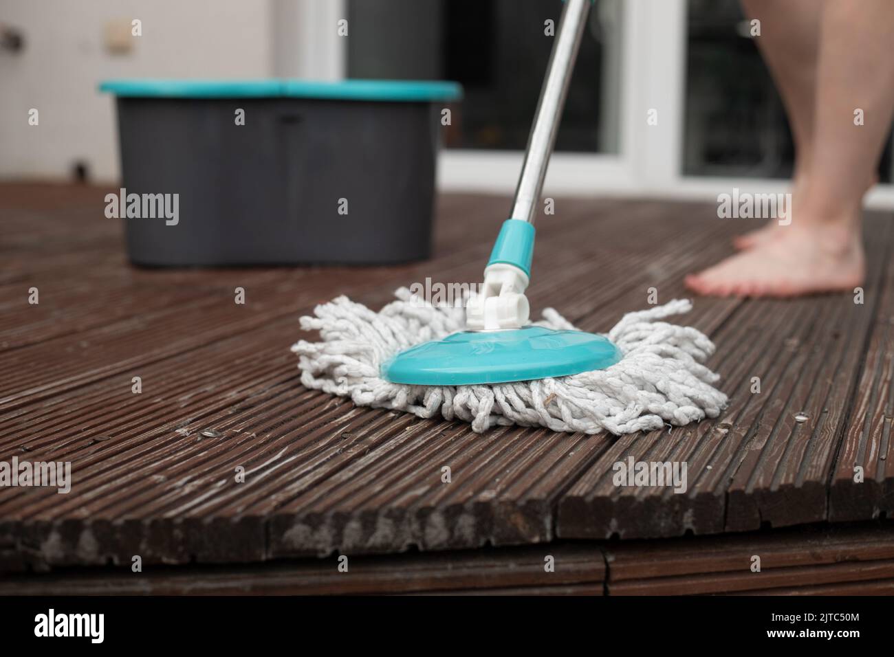 Beschnittenes Foto von Beinen einer jungen Frau, die den braunen Holzboden der Veranda mit einem Spinnmopp in der Nähe eines grauen Plastikeiers putzte. Stockfoto