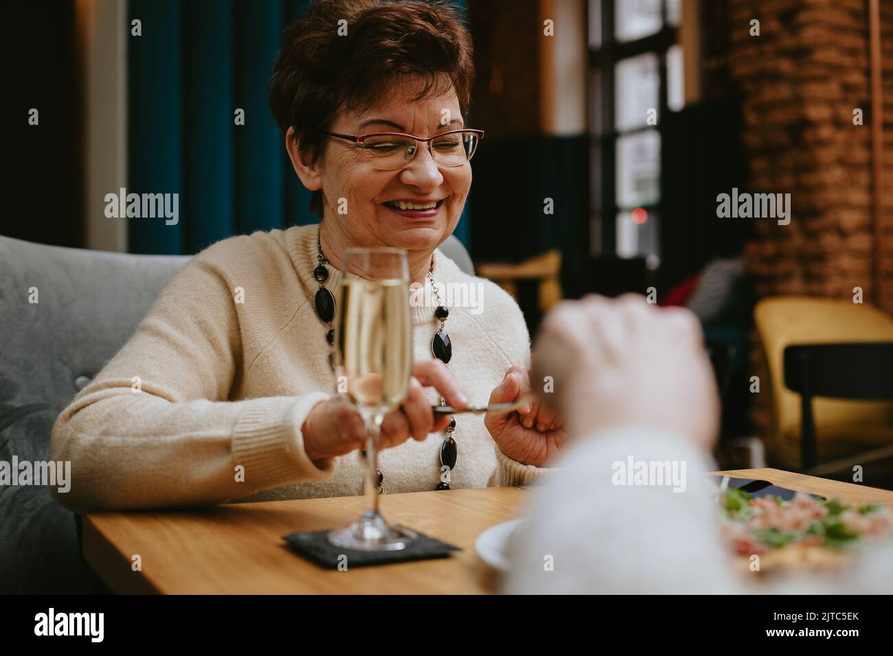 Freunde treffen sich und essen im Restaurant, alte, nette dunkelhaarige Dame mit einem Lächeln essen am Tisch im Cafe-Saal. Alte Freunde trinken Champagner Stockfoto