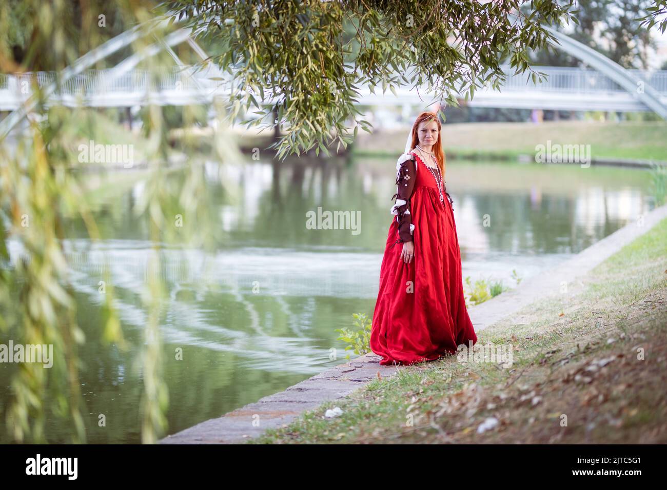 Rothaarige schlanke Mädchen in einem roten Kleid in der venezianischen Mode des 15.. Jahrhunderts posiert vor einem kleinen Teich, voller Länge Porträt Stockfoto