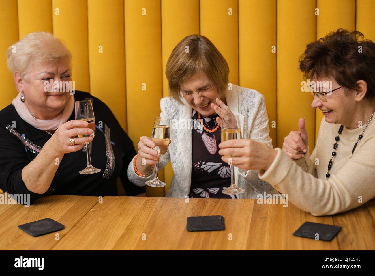 Drei ältere Frauen, alte Freunde treffen sich im Restaurant. Rentner, die am Tisch in einem Zimmer mit gelben Wänden sitzen und fröhlich Champagner trinken Stockfoto