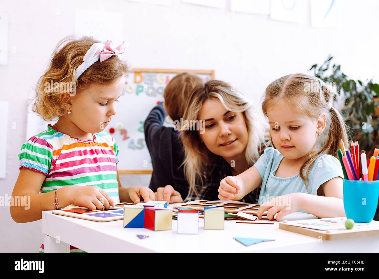 Schöne kleine Mädchen mit Interesse falten farbige Würfel auf dem Schreibtisch im Spielzimmer des Kindergartens. Junger Erzieher, der mit Kindern am Tisch auf dem Boden sitzt Stockfoto