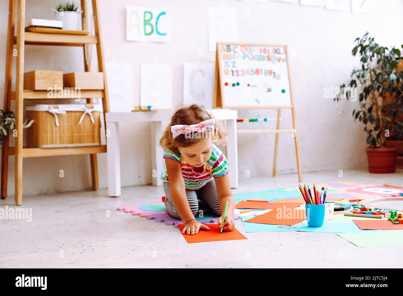 Hübsches kleines Mädchen, das auf den Knien auf dem Boden im Spielzimmer sitzt und ein Bild auf einem roten Blatt Papier mit Marker zeichnet. Kindergartener entwickeln künstlerisch Stockfoto