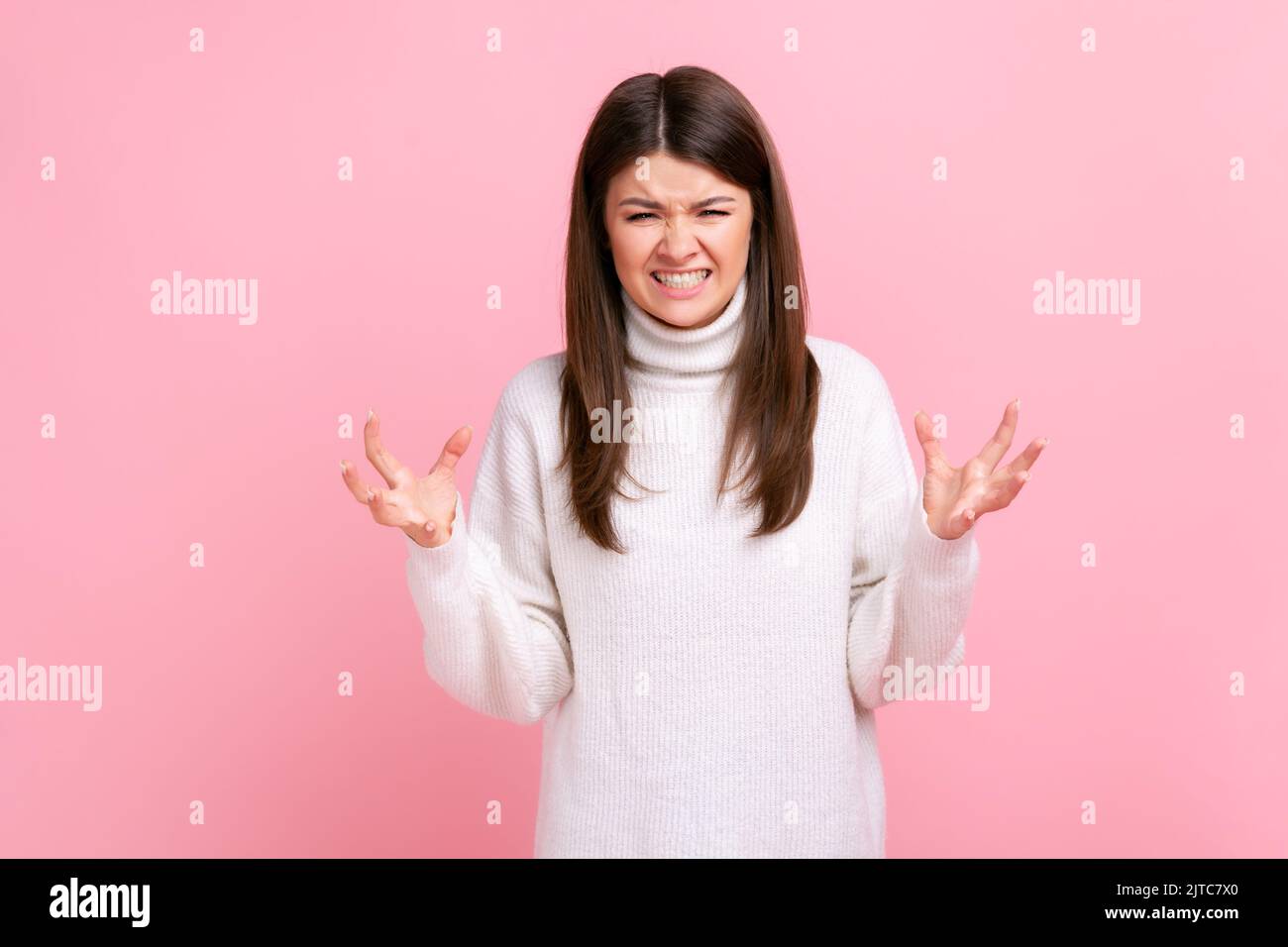 Porträt einer wütenden Frau, die mit erhobenen Armen steht und Aggression, Schreien, Stirnrunzeln ausdrückt und einen weißen Pullover im lässigen Stil trägt. Innenaufnahme des Studios isoliert auf rosa Hintergrund. Stockfoto
