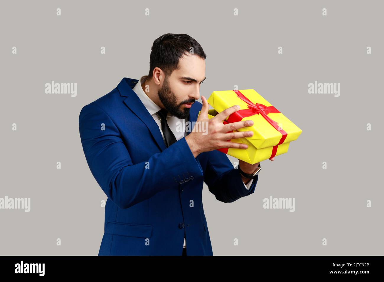 Schöner bärtiger Mann, der Geschenk auspackte, in den Kasten schaute mit neugierigem ernsten Ausdruck, Geburtstagsständchen, Anzug im offiziellen Stil tragend. Innenaufnahme des Studios isoliert auf grauem Hintergrund. Stockfoto