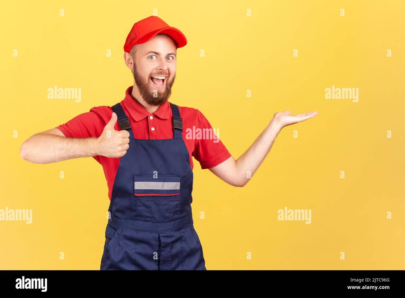 Porträt eines optimistischen Handwerkers, der auf seiner Handfläche einen Platz für Werbematerial vorstellt, den Daumen nach oben zeigt, Overalls trägt und eine rote Mütze trägt Innenaufnahme des Studios auf gelbem Hintergrund. Stockfoto