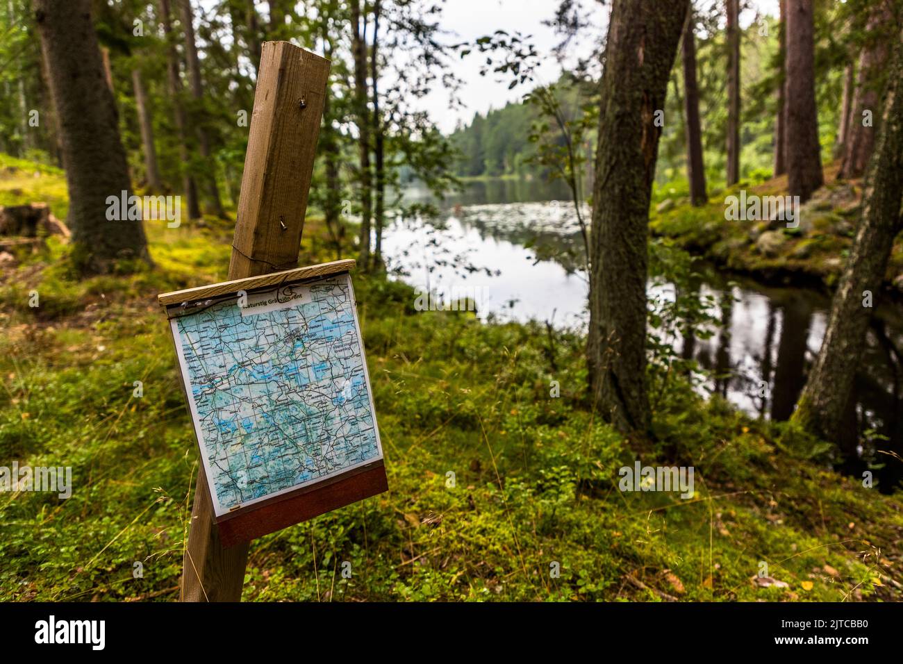 Eine Wanderkarte an einem Holzpfosten zeigt das Gebiet um einen See in der Nähe von Sliparbol, Gemeinde Vingåker, Schweden. Moosbedeckte Kiefern säumen das Ufer. Ein kleiner Bach fließt rechts in den See. Stockfoto