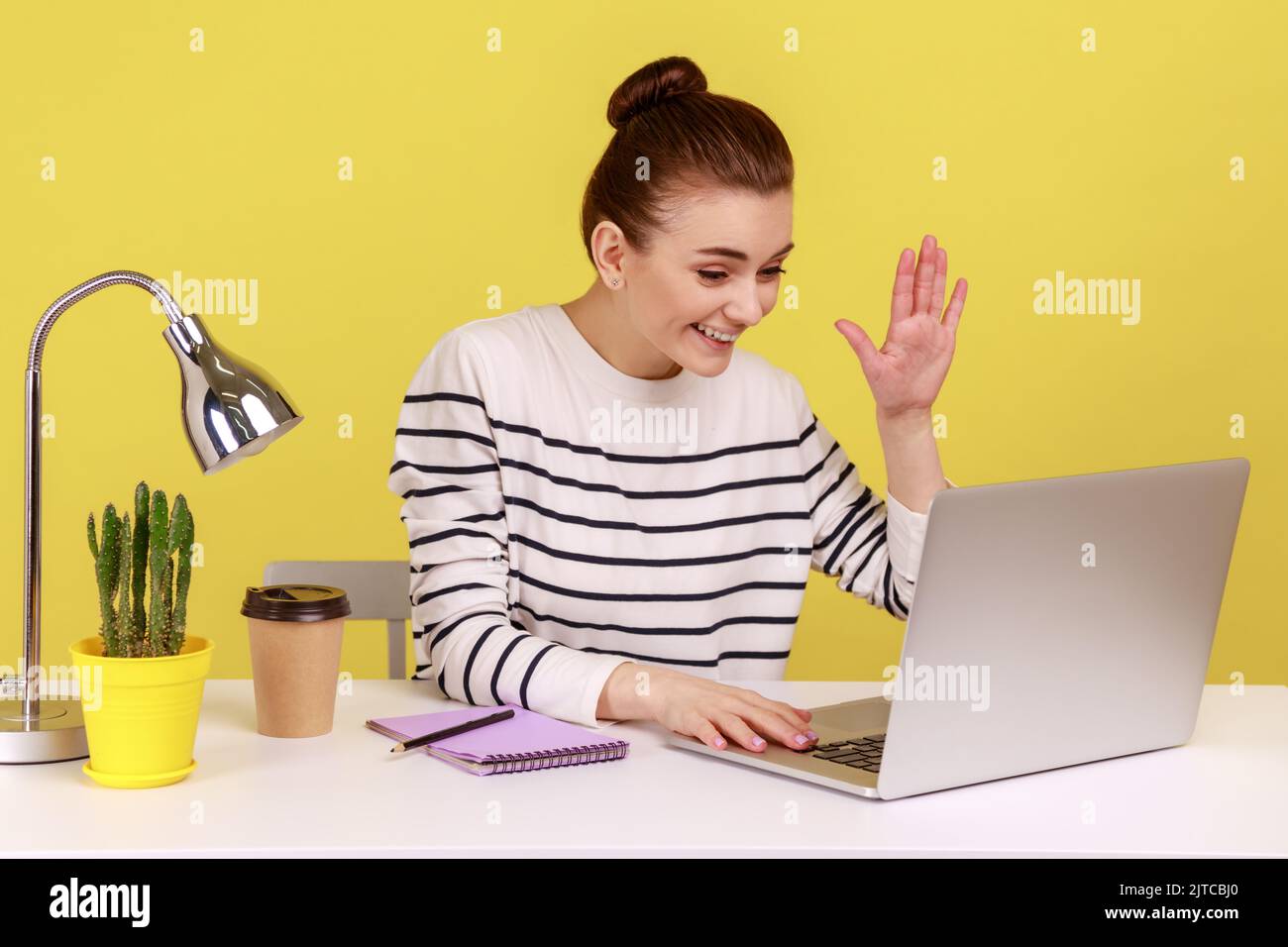 Freundliche Büromanagerin winkt Hand auf den Laptop-Bildschirm, spricht mit einem Freund in einem Videoanruf, Online-Konferenz im Heimbüro. Studio-Innenaufnahme isoliert auf gelbem Hintergrund. Stockfoto