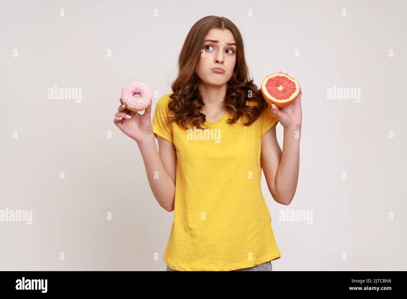 Gesunde Früchte im Vergleich zu kalorienreicher Nachspeise. Porträt eines verwirrten, lockigen Teenagers im lässigen T-Shirt, das zwischen Grapefruit und Donut wählen kann. Innenaufnahme des Studios isoliert auf grauem Hintergrund. Stockfoto