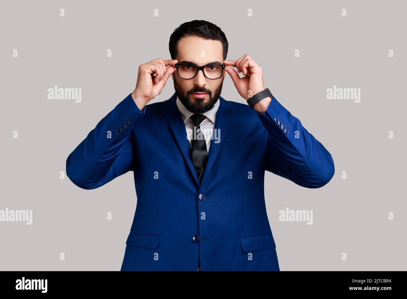 Porträt eines ernsthaften bärtigen Geschäftsmannes, der die Brille ablegt und die Kamera mit herrischem Ausdruck anschaut und einen Anzug im offiziellen Stil trägt. Innenaufnahme des Studios isoliert auf grauem Hintergrund. Stockfoto