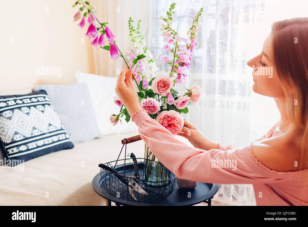 Frau macht Strauß von Rosen und Füchshandschuhen Blumen in Vase auf dem Tisch zu Hause. Einrichtung und Einrichtung im Sommer. Stockfoto