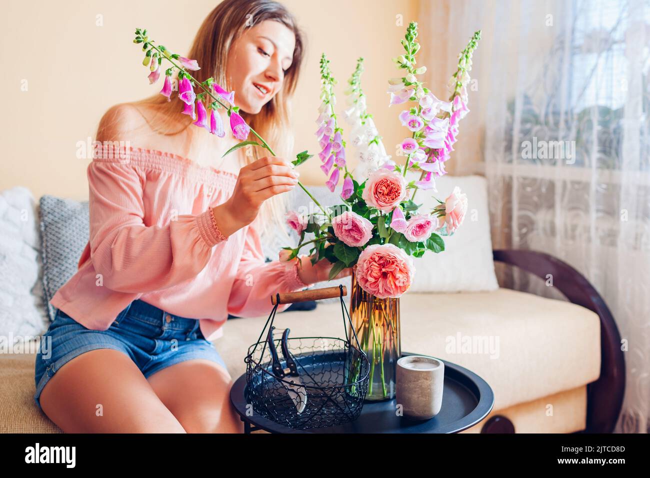 Frau macht Strauß von Rosen und Füchshandschuhen Blumen in Korb in Vase zu Hause gepflückt. Einrichtung und Einrichtung im Sommer. Stockfoto