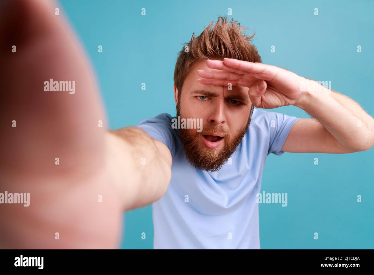 Porträt eines jungen erwachsenen, hübschen, neugierigen bärtigen Mannes, der mit aufmerksamer Sicht und Blick in die Ferne blickt und die Hand nahe an der Stirn hält. Innenaufnahme des Studios isoliert auf blauem Hintergrund. Stockfoto