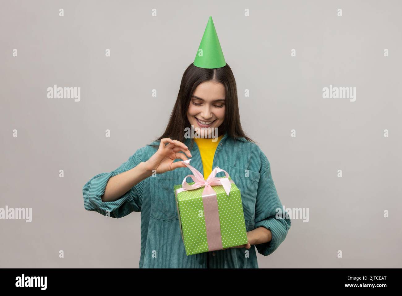 Porträt von lächelnden Frau Partei Kegel Hut Eröffnung Geburtstagsgeschenk auf Party-Feier, ziehen Band, glücklich, tragen lässigen Stil Jacke. Innenaufnahme des Studios isoliert auf grauem Hintergrund. Stockfoto