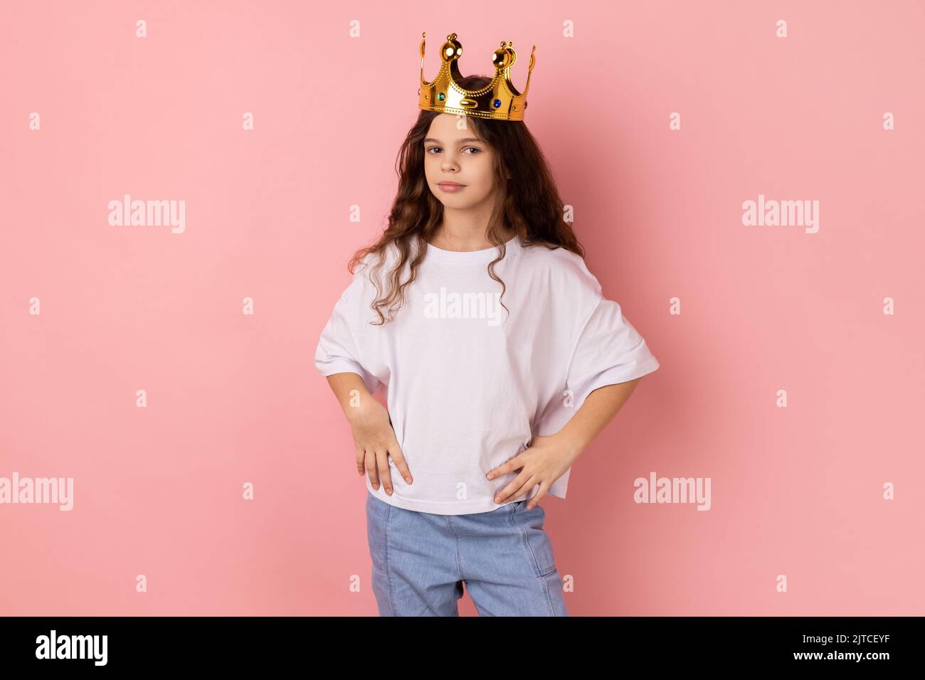 Porträt eines kleinen Mädchens in weißem T-Shirt, das egoistisch auf die Kamera schaut, die mit Krone auf dem Kopf posiert, vorgibt, Prinzessin zu sein, die Hände an den Hüften hält. Innenaufnahme des Studios isoliert auf rosa Hintergrund Stockfoto