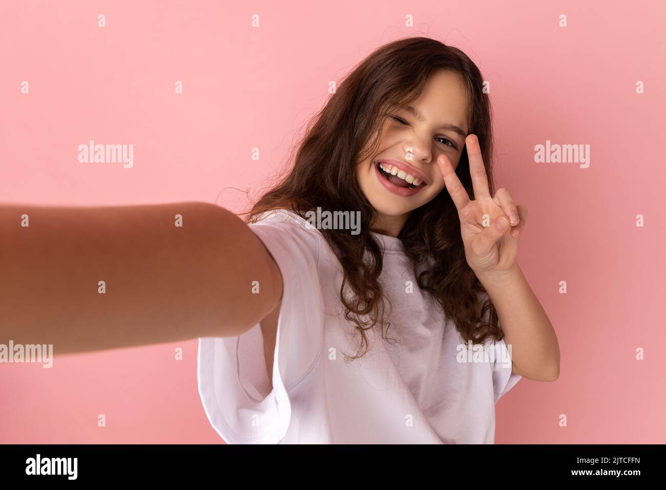 Porträt eines optimistischen kleinen Mädchens in weißem T-Shirt, das Selfie macht, auf den POV der Kamera schaut, aus der Perspektive des Fotos, mit einer Siegesgeste. Innenaufnahme des Studios isoliert auf rosa Hintergrund. Stockfoto