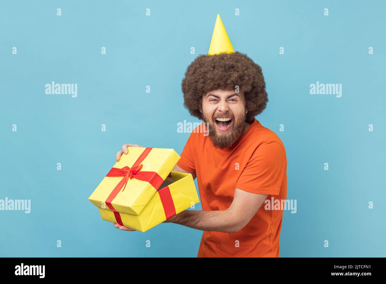 Porträt eines glücklichen Mannes mit Afro-Frisur in orangefarbenem T-Shirt und Party-Kegel, ausgepackter Geschenk-Box, Blick auf die Kamera und Lachen. Innenaufnahme des Studios isoliert auf blauem Hintergrund. Stockfoto