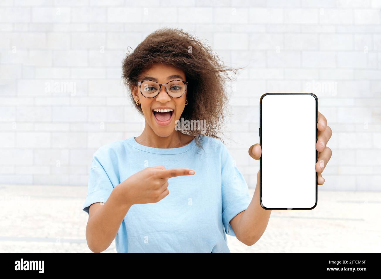 Aufgeregt schockiert staunte afroamerikanische junge Frau mit lockigen Haaren, stilvoll gekleidet, im Freien stehen, zeigt Smartphone mit leerem weißen Mock-up-Bildschirm, zeigt den Finger darauf, schaut überrascht auf Kamera Stockfoto