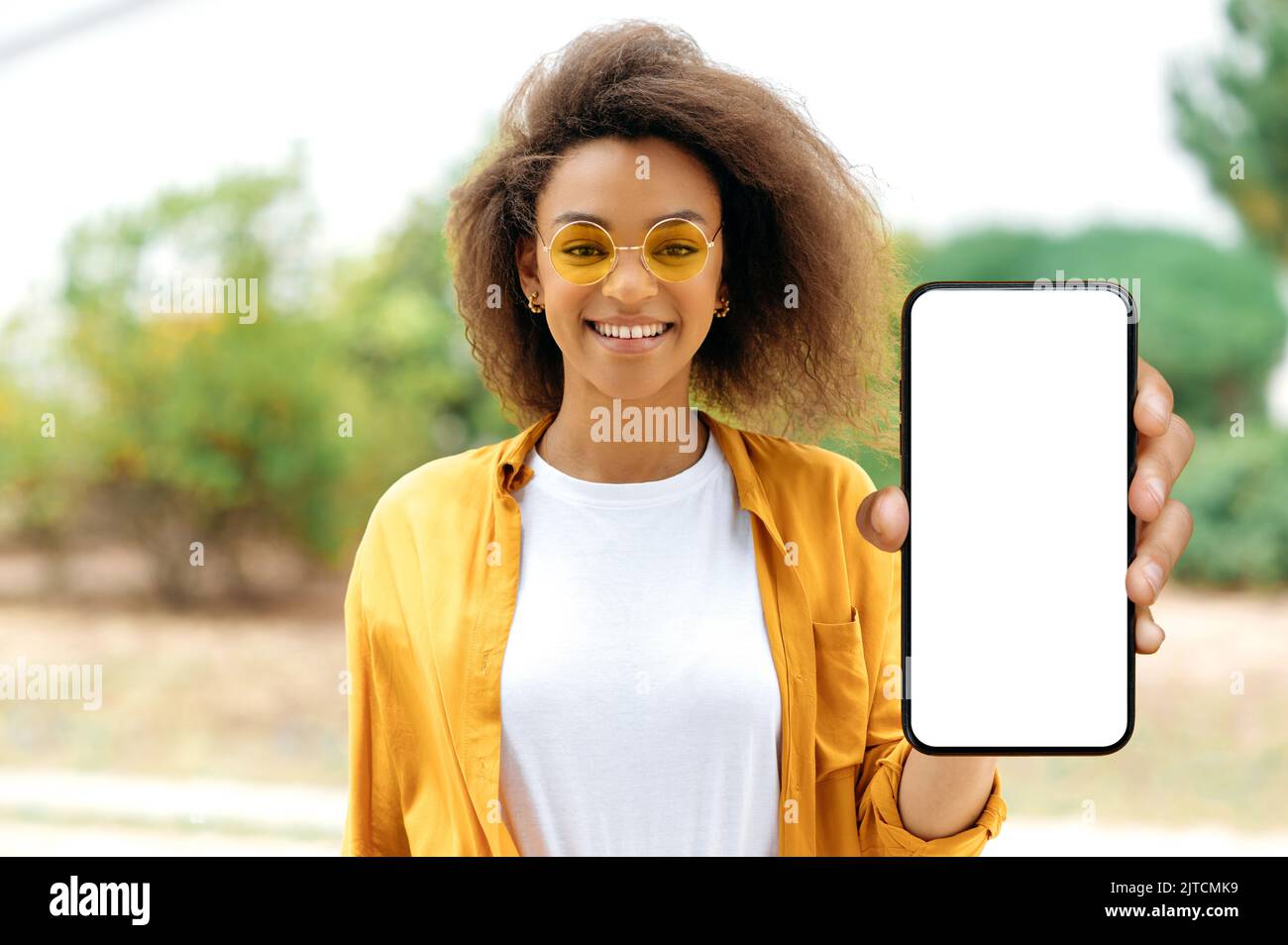 Positive afroamerikanische lockige junge Frau mit fröhlichem Gesichtsausdruck, in lässiger Kleidung, mit oranger Brille, steht im Freien, zeigt Smartphone-Bildschirm mit weißem Mock-up-Bildschirm, schaut auf die Kamera, lächelt Stockfoto