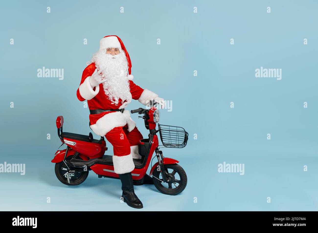 Der weihnachtsmann, der hinter dem Steuer eines roten Elektromopeds sitzt, macht mit seiner Hand eine Horngeste und schaut isoliert auf einem blauen Blick auf die Kamera Stockfoto