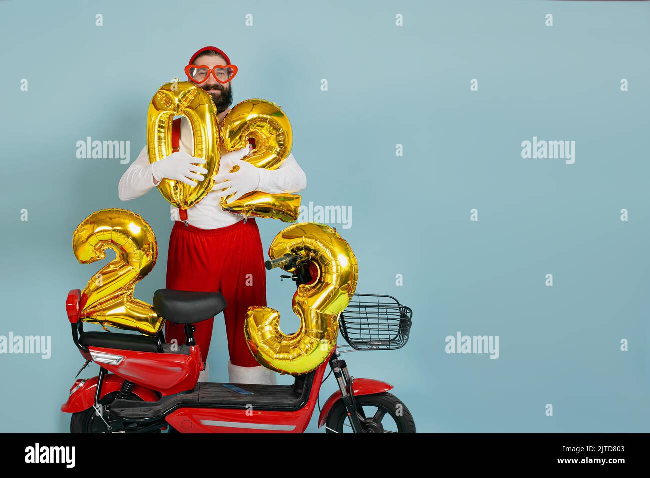 Der junge Erwachsene Hipster-Mann in einem weihnachtsmann-Kostüm und einer großen herzförmigen Brille hält aufblasbare Zahlen 2023 in den Händen auf einem roten Elektro-Scooter auf einem blauen Stockfoto