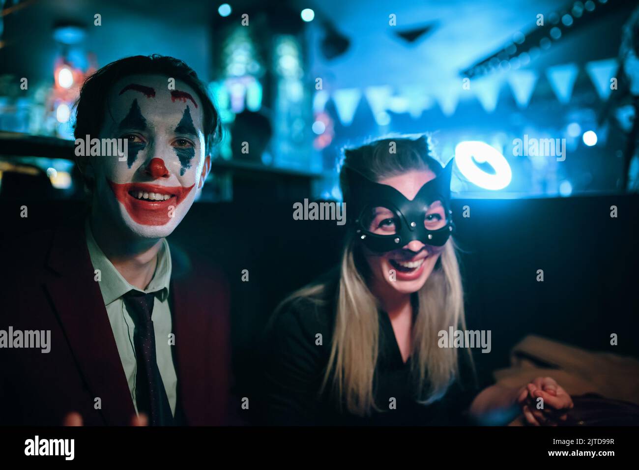 Frau in Katzenmaske und Kerl mit Joker Make-up sitzt am Tisch in der Kneipe. Stockfoto