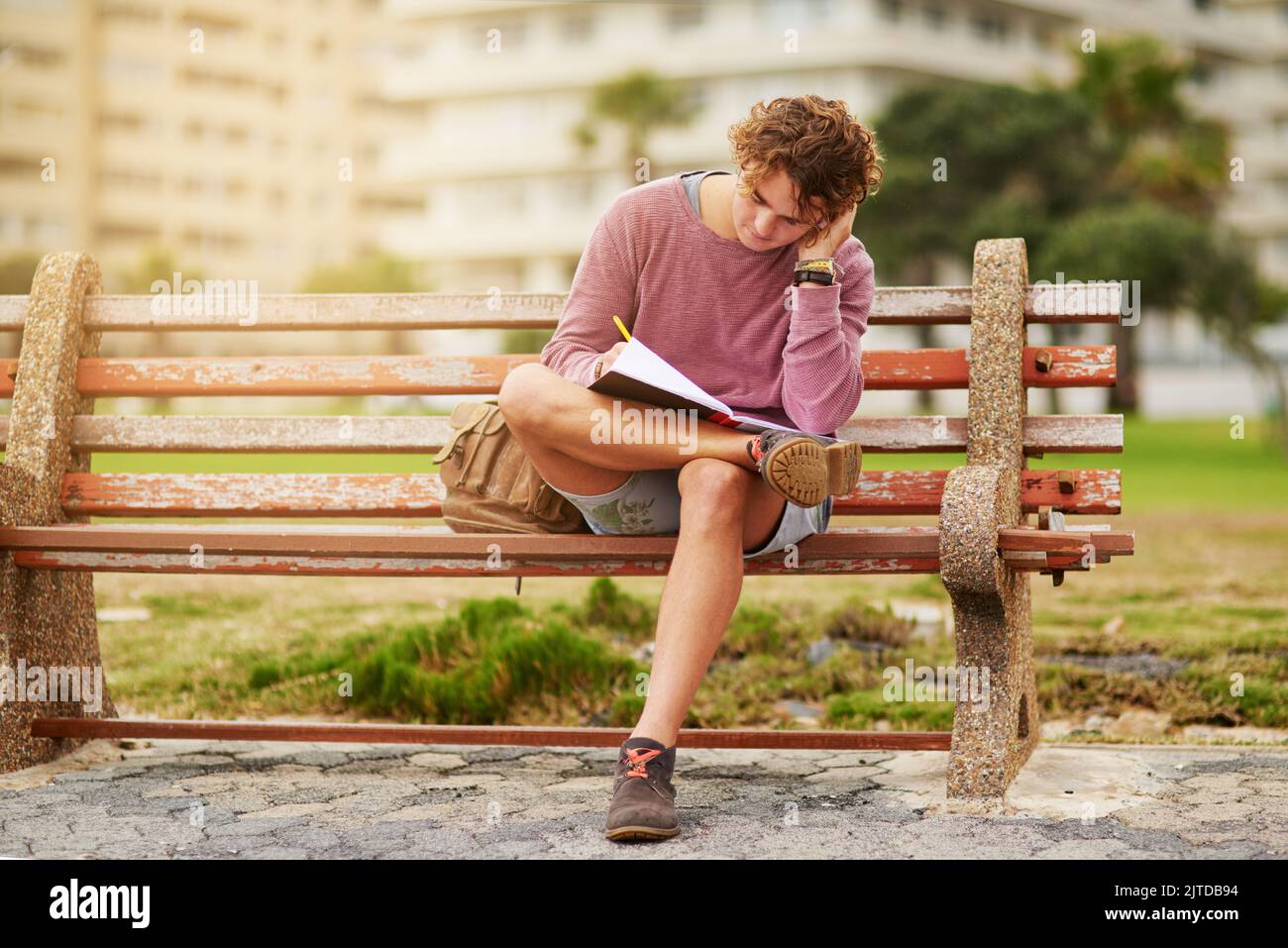 Ein junger Mann, der auf einer Parkbank sitzt und in einem Notizbuch schreibt. Stockfoto
