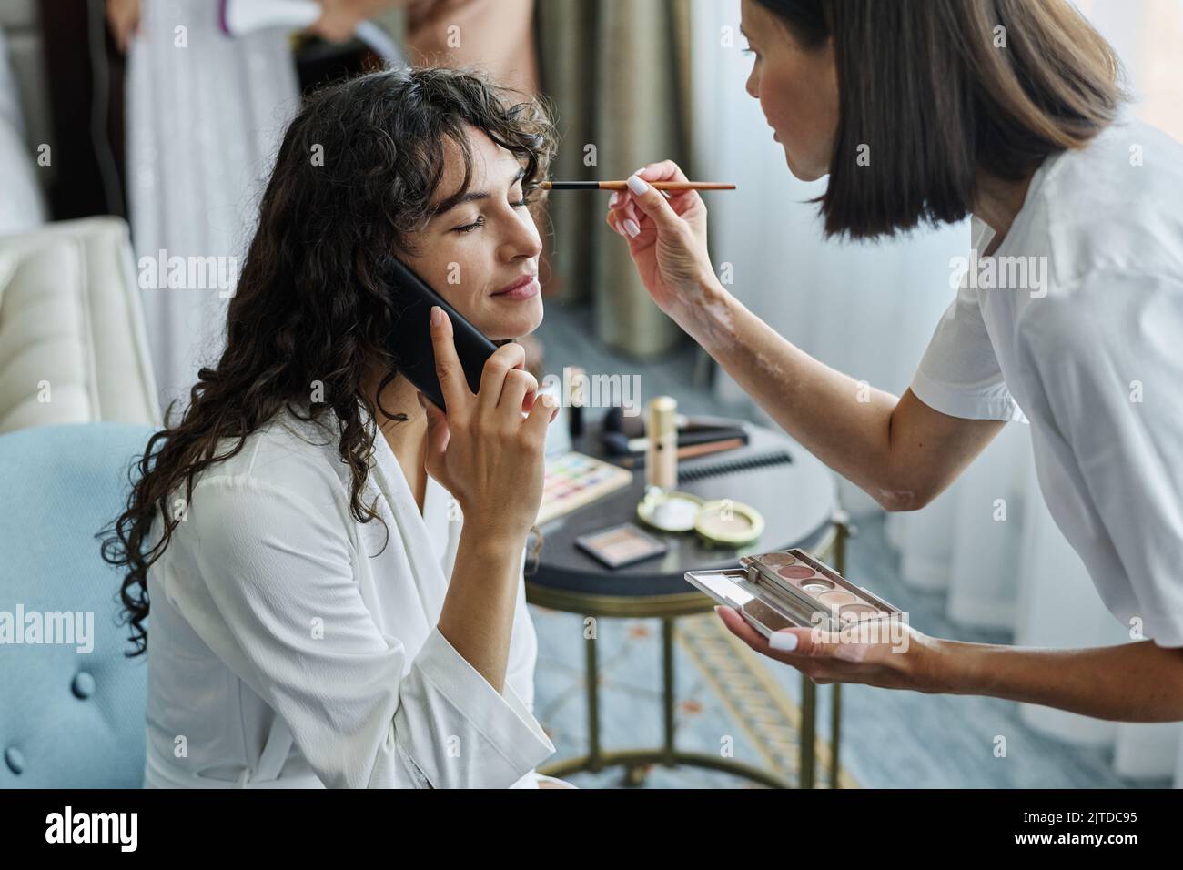 Junge lächelnde Brünette Frau im weißen Bademantel im Gespräch auf dem Handy, während Make-up-Künstler Lidschatten auf ihre Augen Stockfoto
