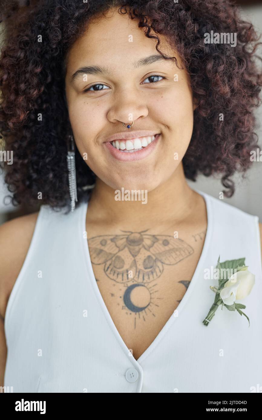 Junge lächelnde schwarze Frau mit Tattoo auf der Brust in weißer Weste mit Knopflochblume, die bei der Hochzeit vor der Kamera steht Stockfoto