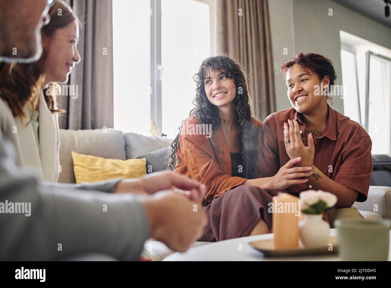 Glückliche junge, multiethnische Frau, die einen Verlobungsring am Finger zeigt, während sie im Wohnzimmer mit den Eltern ihrer Freundin spricht Stockfoto