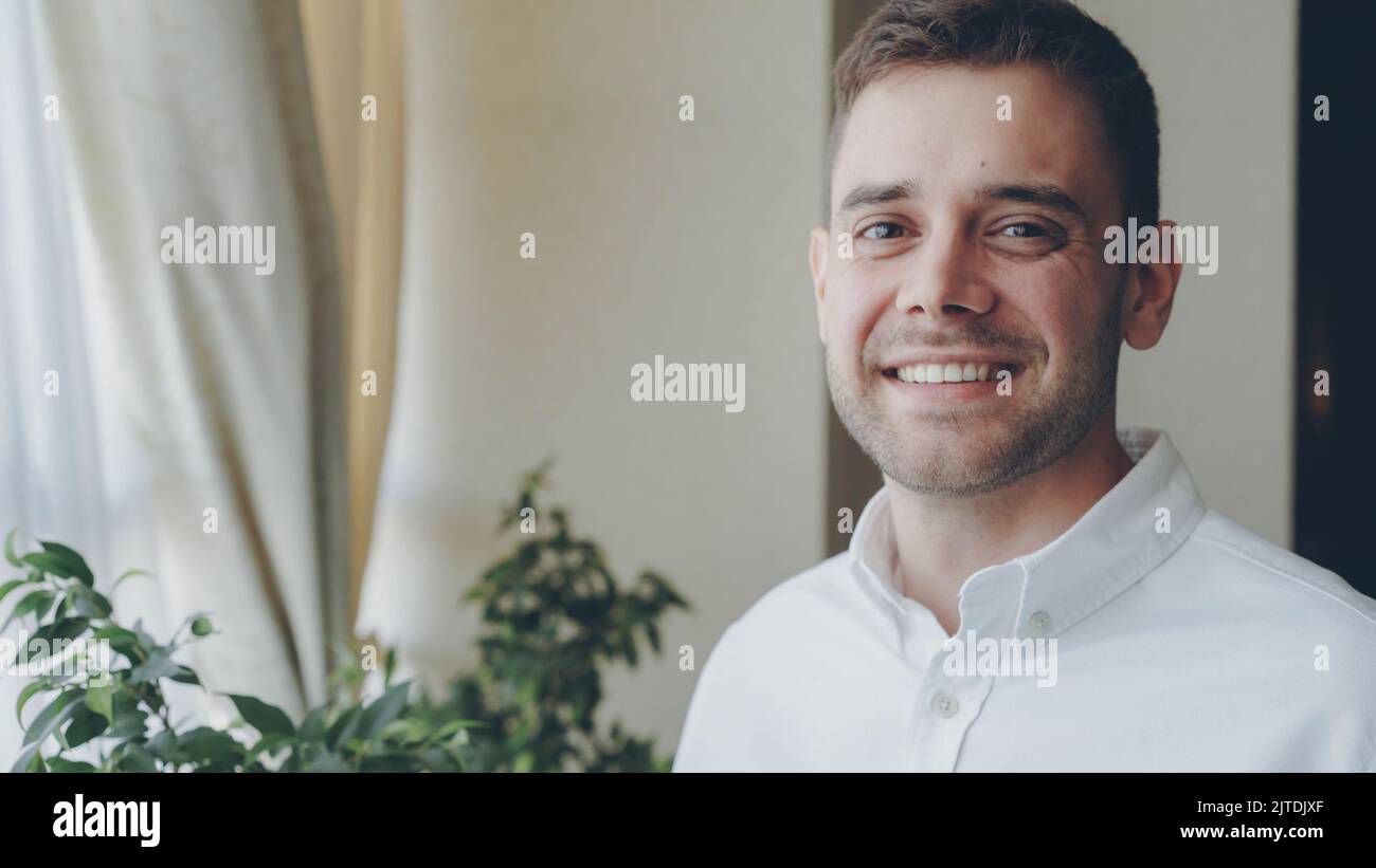 Nahaufnahme eines dunkelhaarigen, hübschen jungen Mannes in weißem Hemd, der fröhlich lächelt und die Kamera anschaut. Junge Menschen, schöne Inneneinrichtung und Glückskonzept. Stockfoto