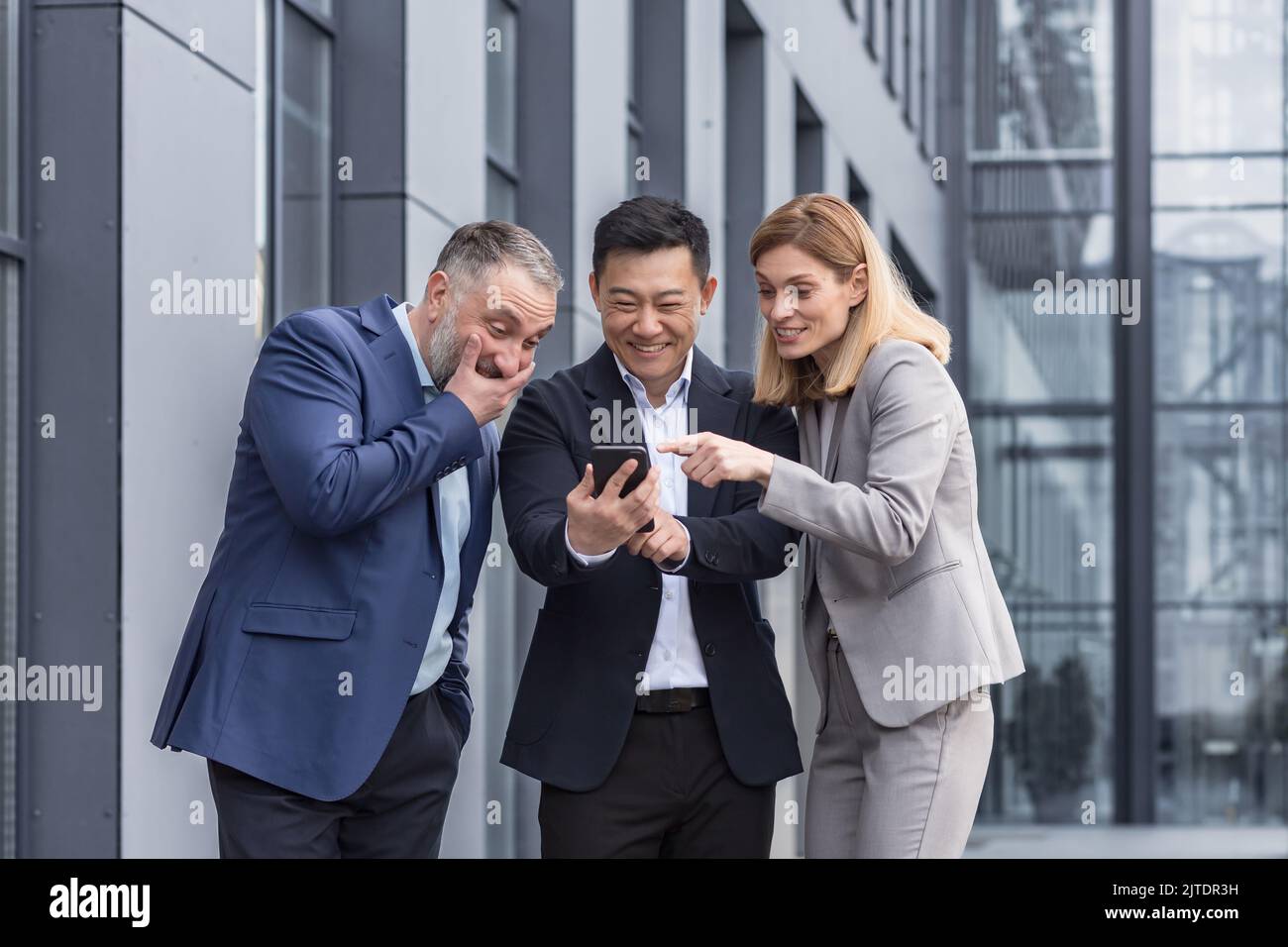 Vielfältige Geschäftsgruppe, drei männliche und weibliche Mitarbeiter vor dem Bürogebäude, fröhlich lächelnd und glücklich beim Ansehen von Videos am Telefon Stockfoto