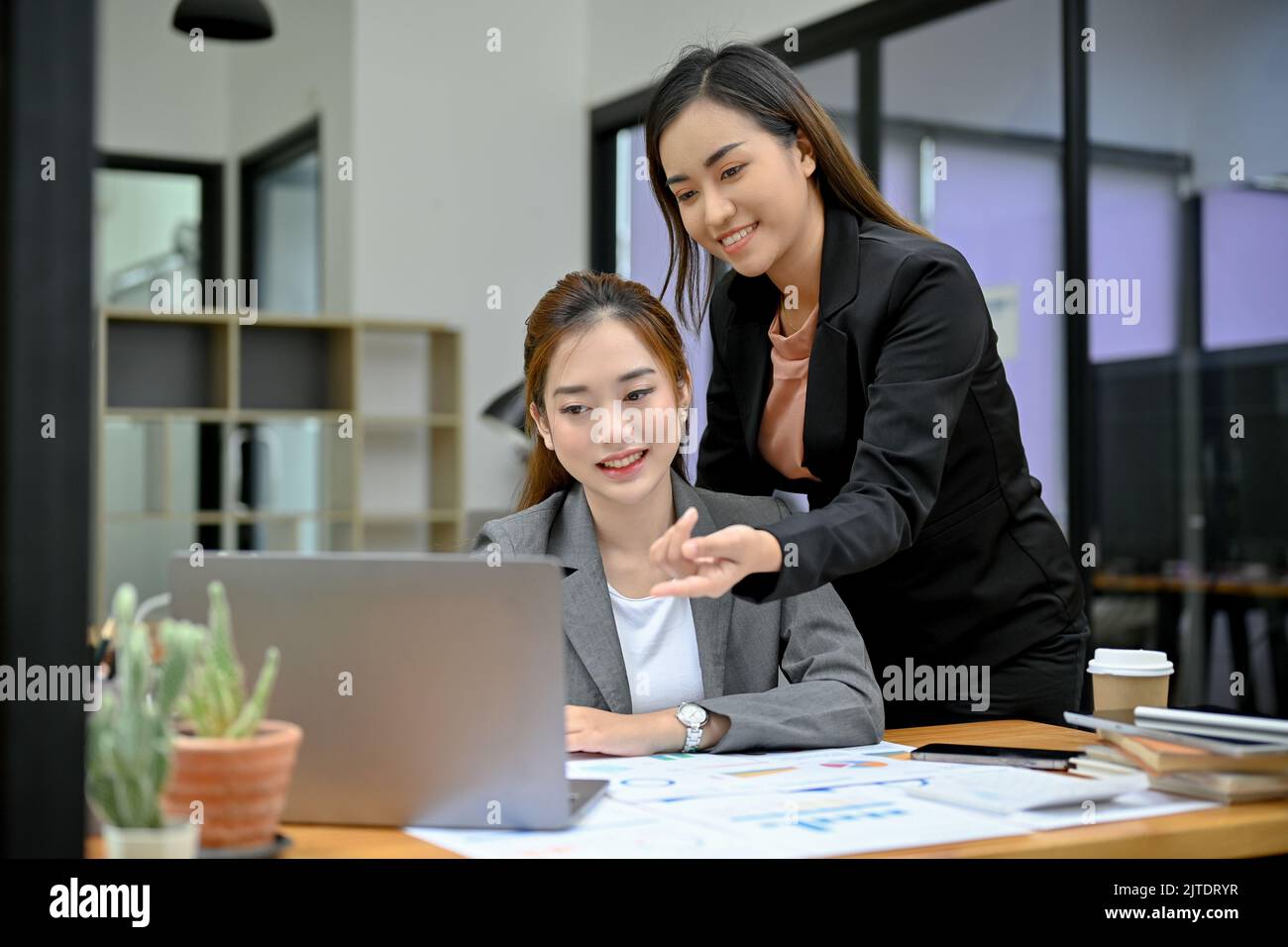 Professionelle und fröhliche asiatische Managerin, die die Arbeit ihrer jungen weiblichen Mitarbeiterin am Laptop coacht, trainiert und berät. Stockfoto