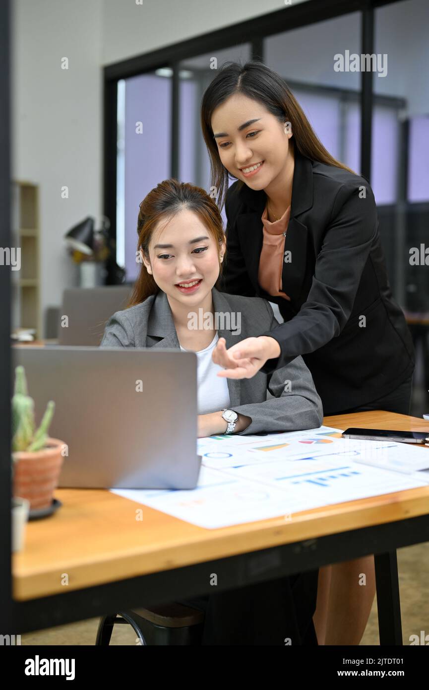 Freundliche und professionelle asiatische Geschäftsfrau oder Managerin, die ihre neueste weibliche Mitarbeiterin bei der Arbeit berät. Teamwork-Konzept Stockfoto