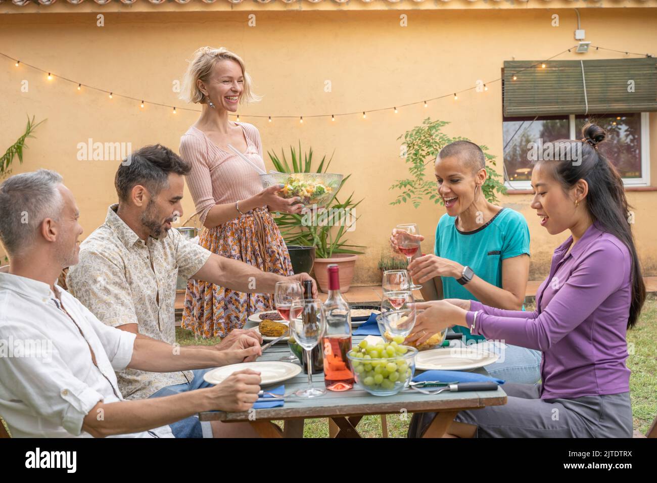 Fröhliche Freunde, die bei einem Sommeressen mit einem Lächeln am Tisch lachen. Die Frau serviert das Essen auf der Terrasse des Hauses. Lächeln, Heiterkeit Konzept. Stockfoto