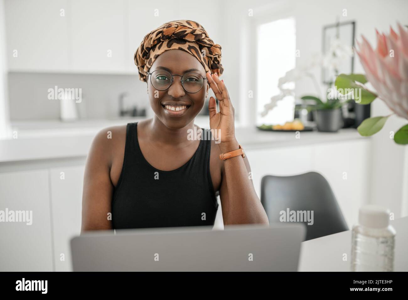 Schwarze Afrikanerin lächelt und hält eine Augenbrille. Blick auf den Laptop, während Sie von zu Hause aus sitzen und arbeiten. Tragen Sie einen traditionellen Kopftuch Stockfoto