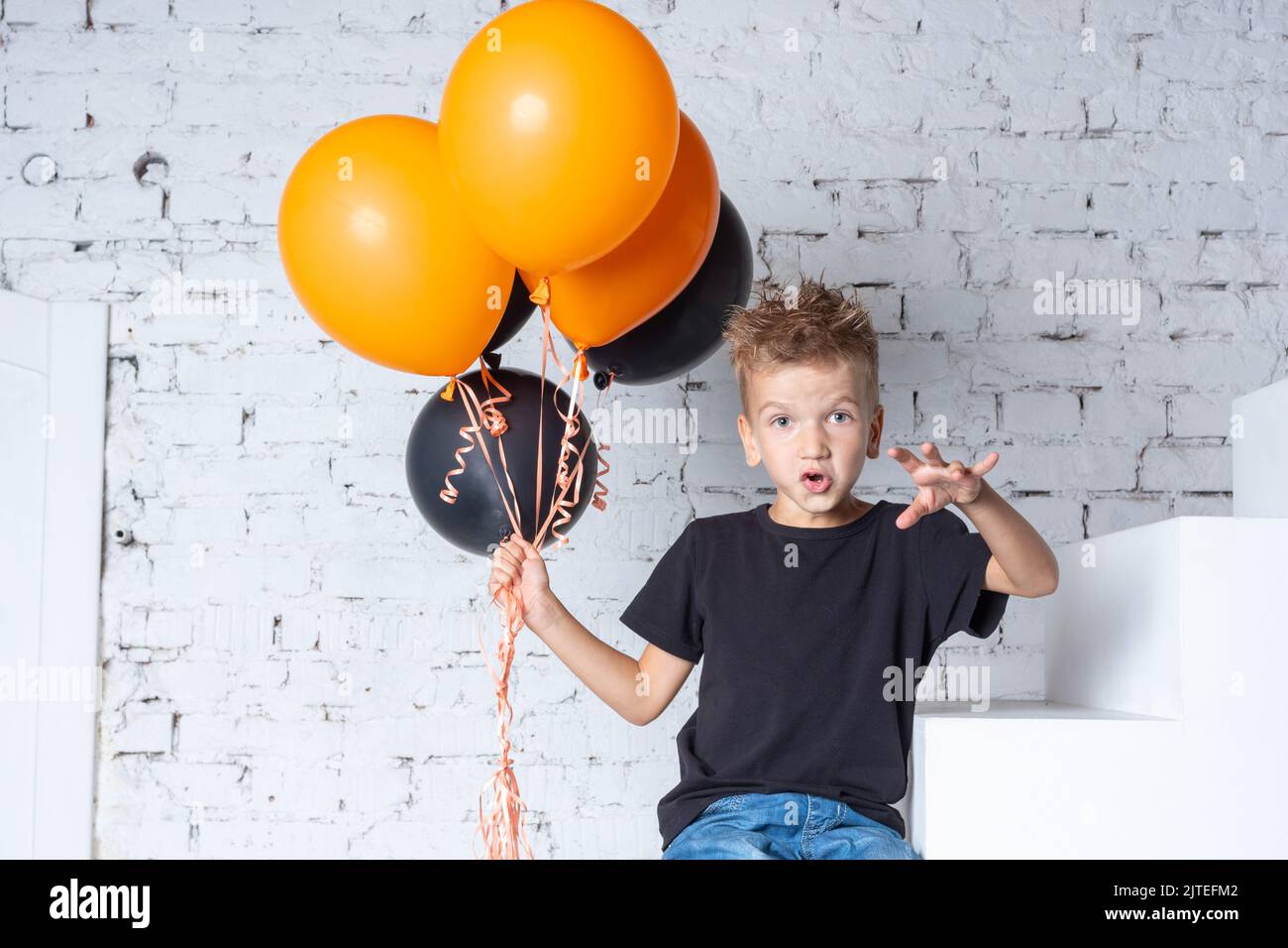 Halloween Kinder. Ein kleiner Junge in einem schwarzen T-Shirt mit orangefarbenen und schwarzen Ballons sitzt auf der Treppe des Hauses. Das Kind macht einen Schrecken Stockfoto