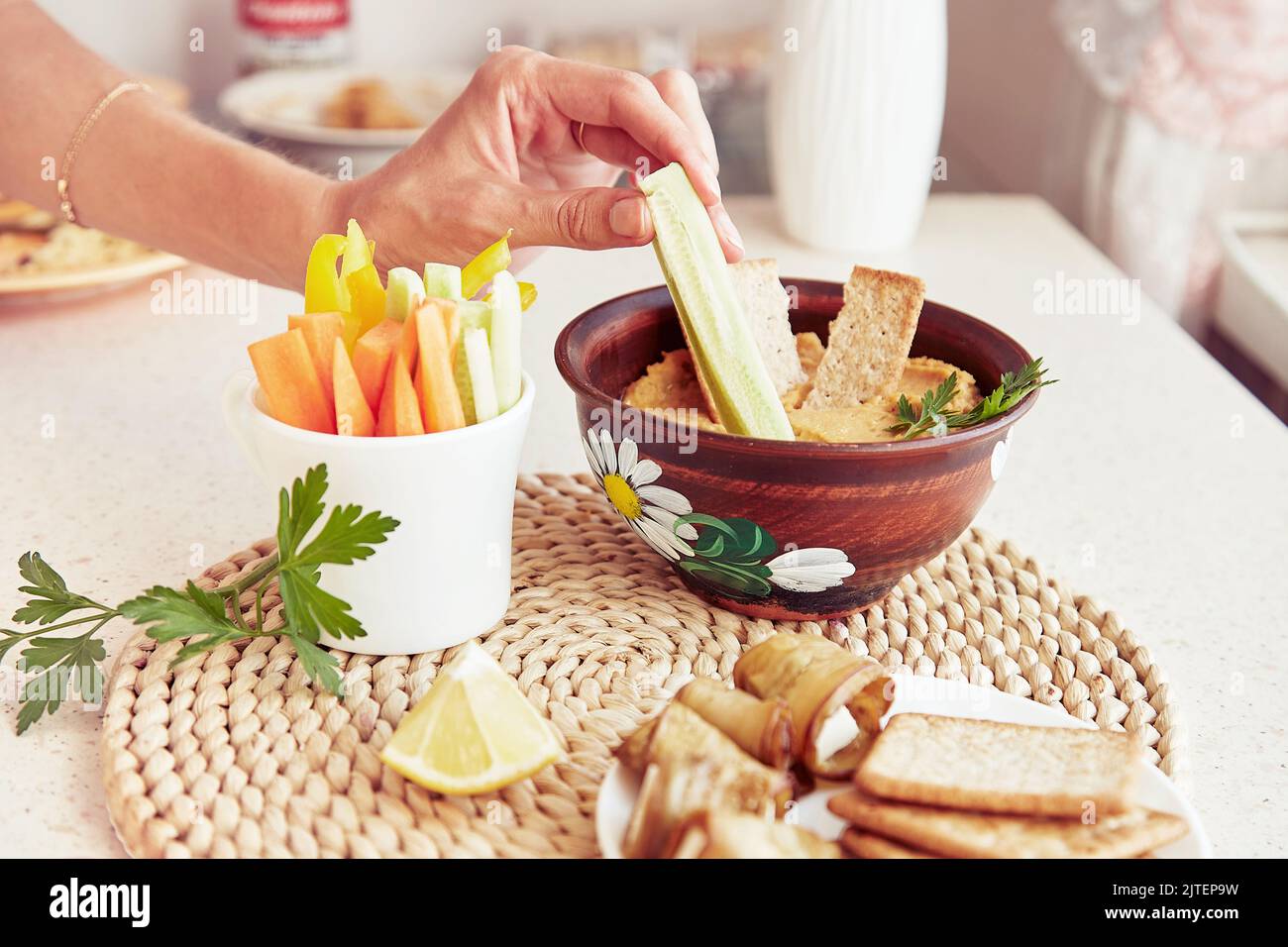 Frau Dip frische Gurke in hausgemachten natürlichen Hummus. Kekse, Petersilie und frisches Gemüse auf dem Tisch. Gesunde vegetarische rustikale Snacks aus nächster Nähe Stockfoto