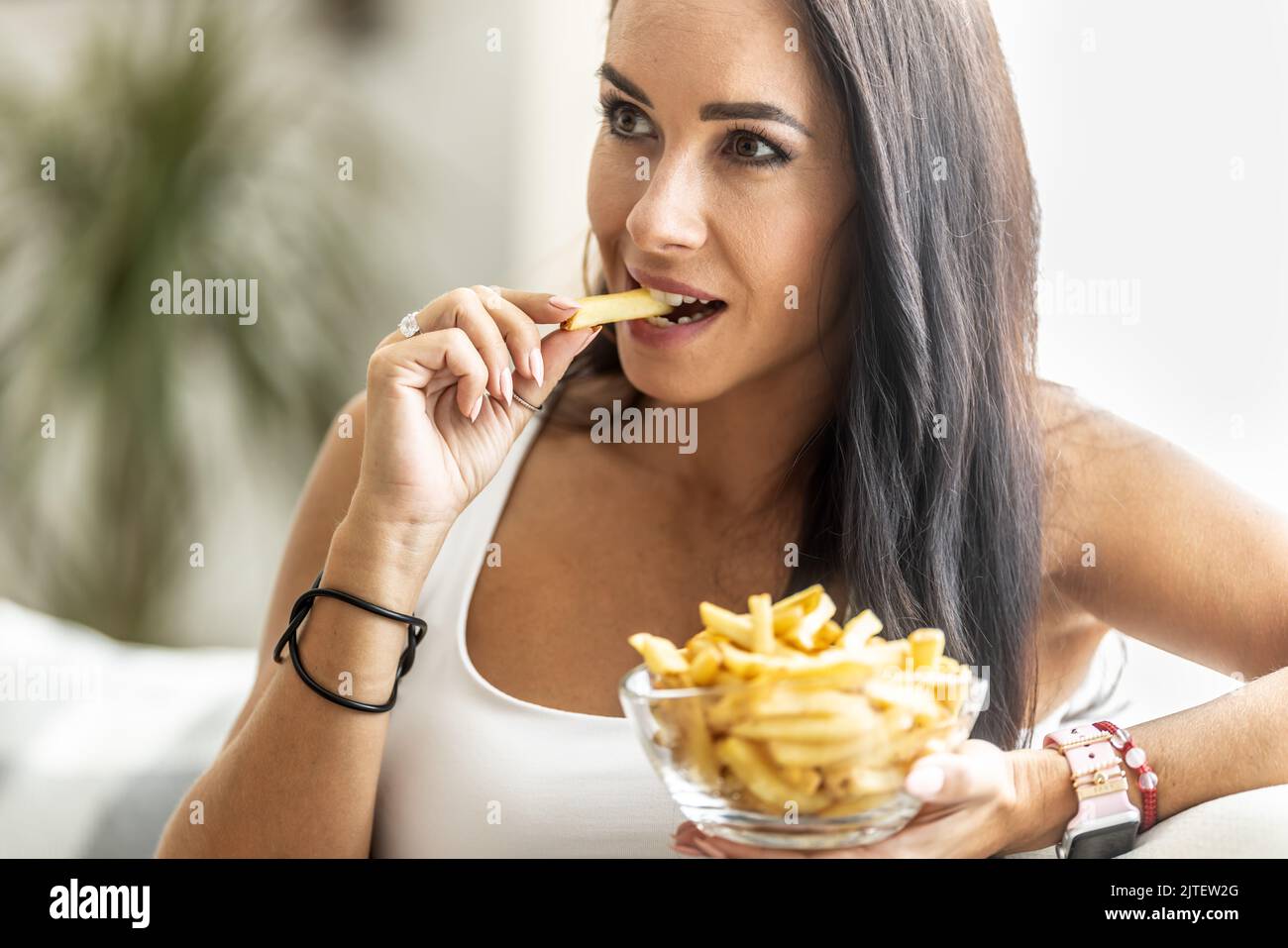 Eine Frau, die eine Schüssel mit Pommes isst, beißt in knusprig gelbe, in den Fingern gebratene französische pommes. Stockfoto
